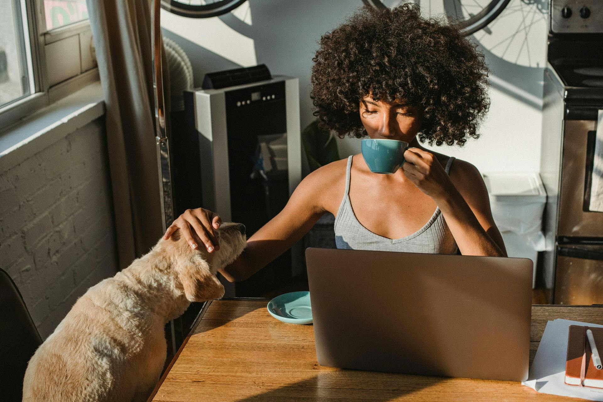 A woman petting her dog while drinking a hot drink and looking at a laptop