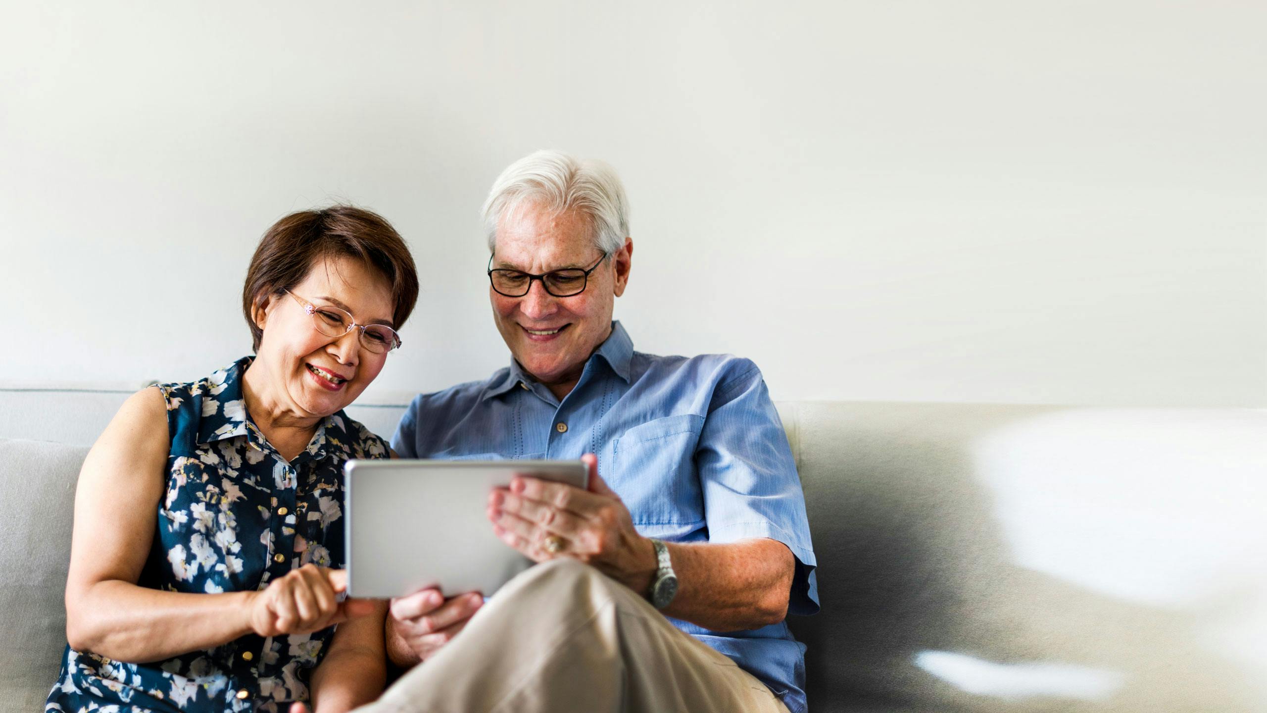 An older couple sitting on the sofa smiling, while looking at a tablet