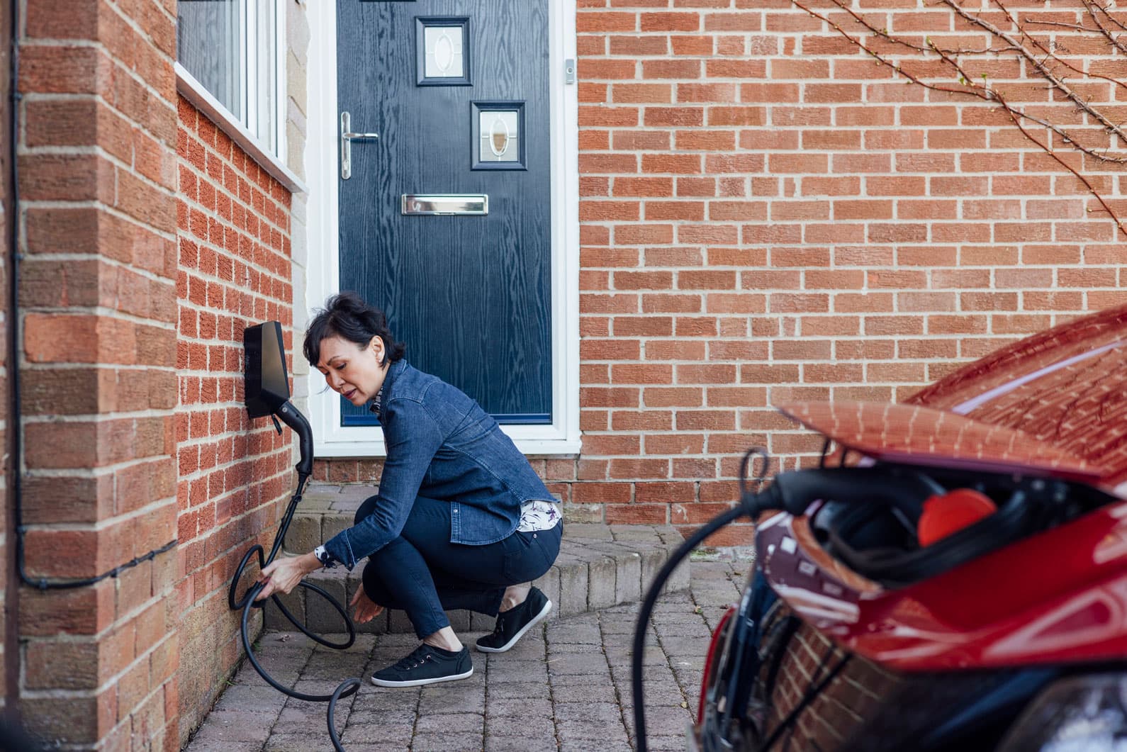 A woman plugging her car into a personal home EV charger outside the front door of a house.