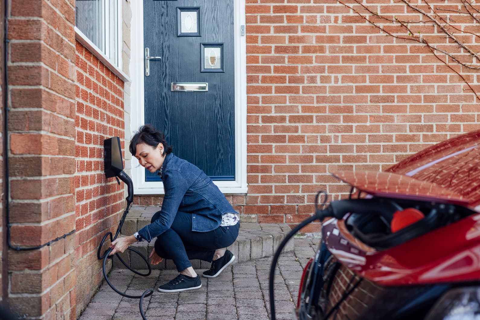 A woman plugging her car into a personal home EV charger outside the front door of a house.