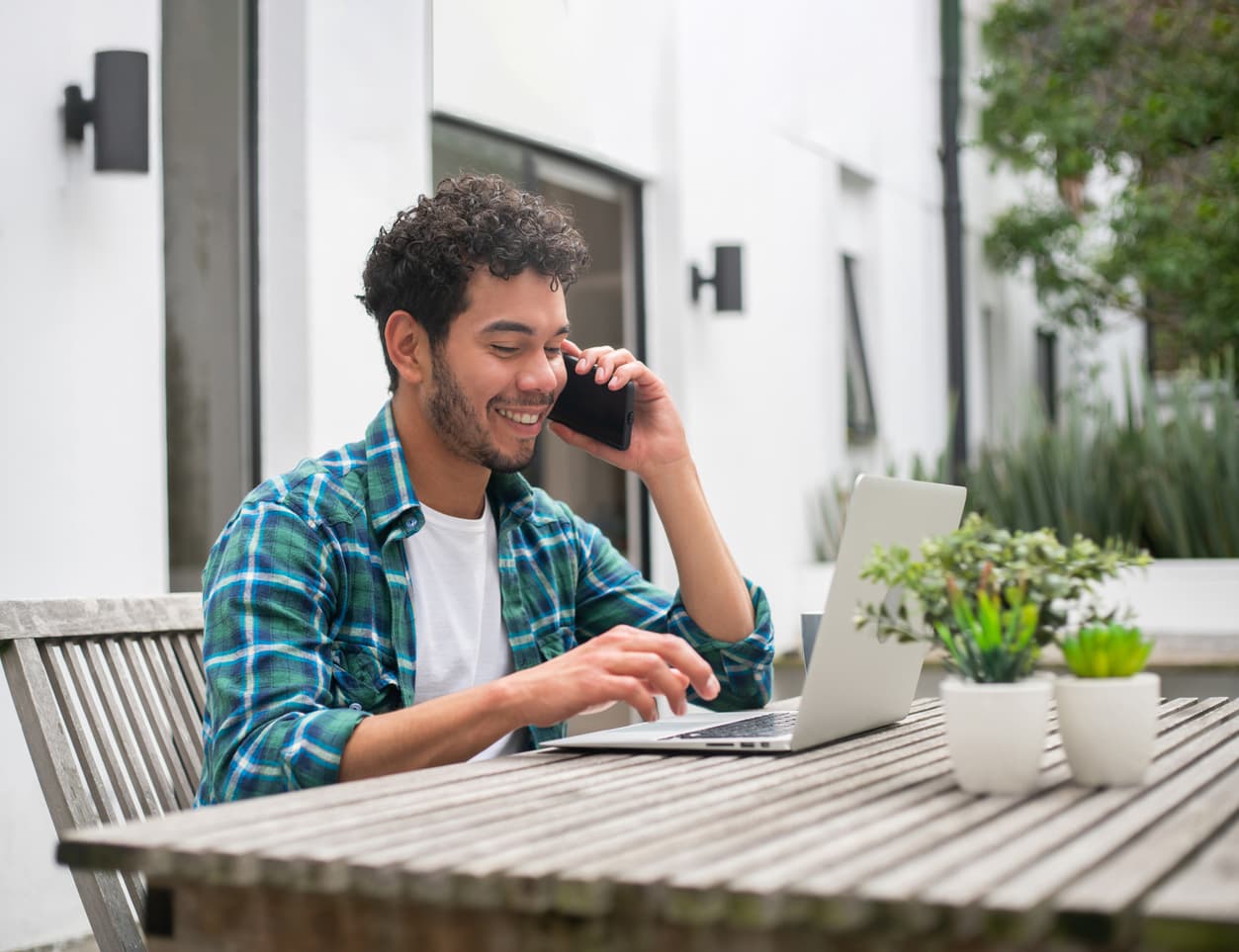A man on the phone in his garden while he scrolls on his laptop.