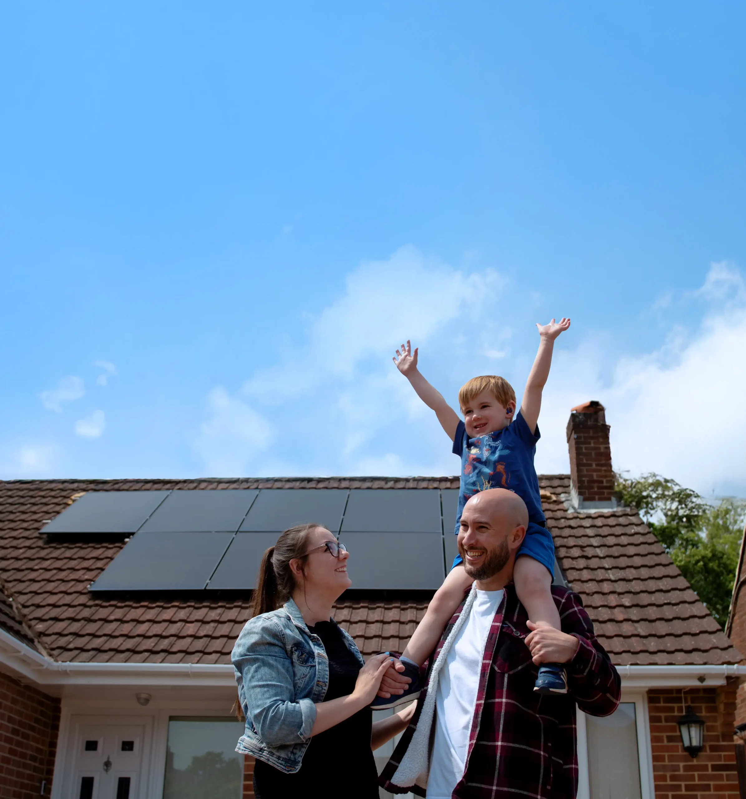A mother & father with their son on his shoulders in front of a property after Utilita Home installed solar panels.